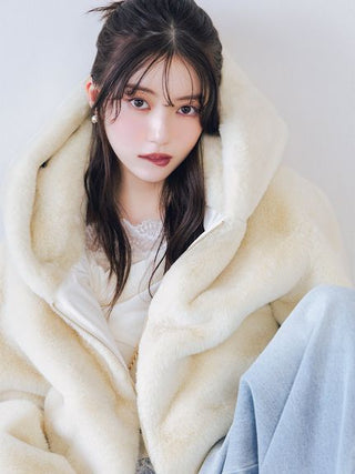 A woman wearing a cream-colored plush faux fur coat over a white top and light blue jeans, posing against a plain light background.