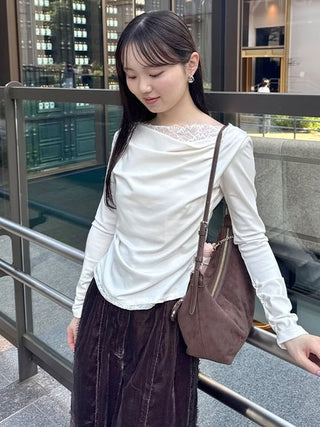 A woman wearing a white long-sleeve top with a lace neckline and a dark brown velvet skirt, carrying a brown shoulder bag in an outdoor urban setting.