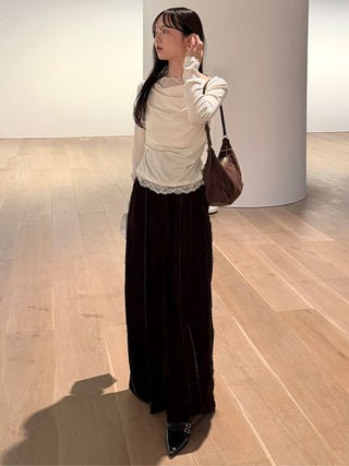 A woman stands in a minimalist gallery or studio with light wood floors and white walls, wearing a cream ruched long-sleeve top with a lace hem, a long dark brown velvet skirt, and black pointed shoes, while carrying a brown shoulder bag.
