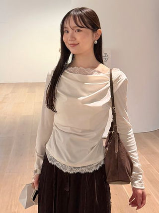 A woman wearing a cream-colored long-sleeve top with lace trim at the neckline and hem, paired with a dark brown skirt and carrying a brown shoulder bag, standing in a minimalist indoor space with light wood floors.