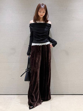A woman models a two-piece outfit consisting of a black off-the-shoulder long-sleeve top with a white lace hem and dark brown wide-leg velvet trousers, holding a black handbag against a textured gray wall.