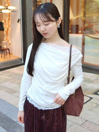 A woman wearing a white long-sleeve top with lace trim and a dark brown skirt, carrying a brown shoulder bag, standing on a city sidewalk.