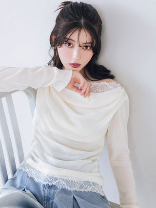 A woman wearing a cream-colored long-sleeve top with lace trim at the neckline and hem, paired with light blue jeans, leaning against a white wall.