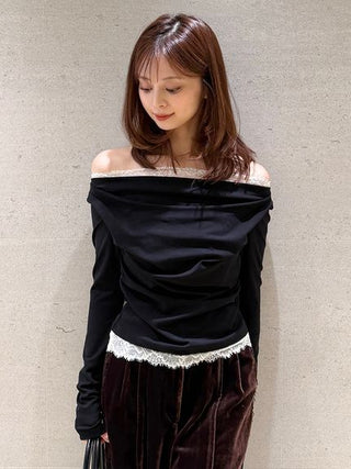 A woman wearing a black off-the-shoulder long-sleeve top with white lace trim and dark brown velvet pants, standing against a textured gray wall.