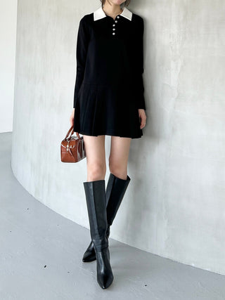 Woman wearing a black dress with white collar and black knee-high boots, standing against a white wall.
