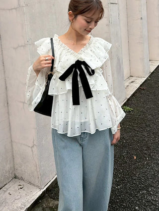 Close-up of a white dotted ribbon frill tunic with layered ruffles, sheer sleeves and a black bow, styled with jeans.