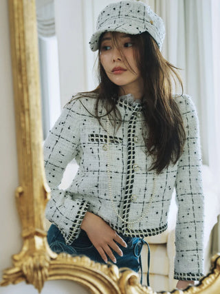 A woman wearing a light gray tweed jacket with black checkered patterns, a matching gray cap, and blue jeans, posing in front of a large ornate gold mirror.