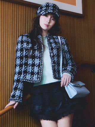 A woman models a black and white tweed houndstooth jacket and matching cap, paired with a black ruffled mini skirt and a light blue handbag, set against a warm wood-paneled background.