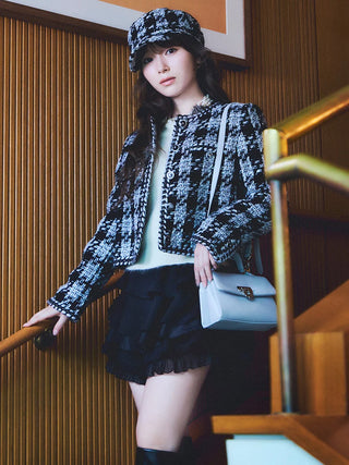 A woman posing on an indoor staircase wearing a black and white patterned tweed jacket, a matching patterned cap, a black ruffled mini skirt, and carrying a light blue shoulder bag.