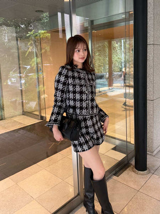 A woman stands in front of a glass building entrance wearing a black-and-white houndstooth tweed mini dress with long sleeves and a peplum hem, paired with black knee-high boots and holding a black handbag.