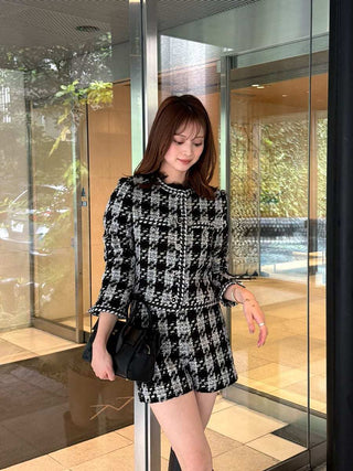 A woman wearing a black and white tweed mini dress with a matching belt, holding a black handbag, standing in front of a glass revolving door.