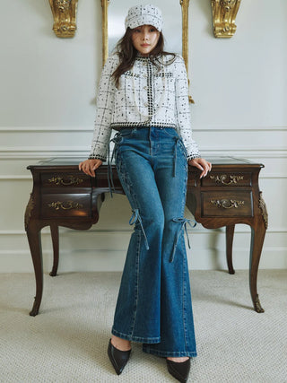 A woman models a white long-sleeve blouse with black polka dots and a black beaded tie-neck detail, paired with high-waisted blue denim flare jeans featuring side tie details, a white knit beanie, and black pointed-toe heels, posed in front of a dark wood writing desk.