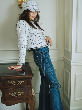 A woman models a white tweed jacket with black grid pattern and matching white tweed cap, paired with blue jeans, leaning against a dark wood cabinet in an elegant room.
