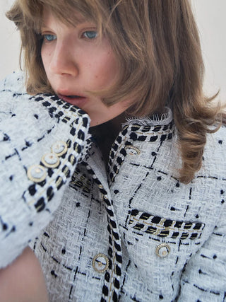 A close-up of a model wearing a black and white tweed jacket with gold buttons and trim, set against a plain light background.