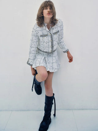 A model wearing a white tweed jacket and matching mini skirt set with black checkered trim, paired with black knee-high boots, posing against a white wall.