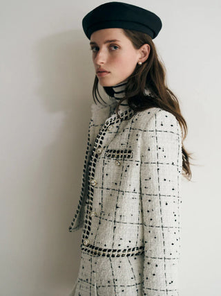 A woman wearing a black beret, a black-and-white tweed jacket with black polka dots and checkered trim, and a black turtleneck, posed against a plain off-white wall.