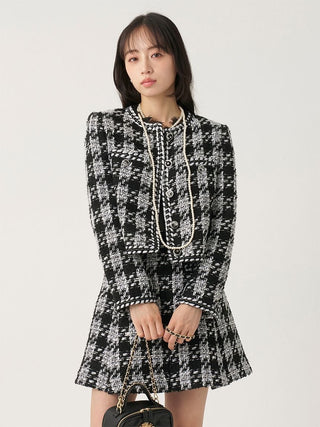 A woman modeling a black and white tweed mini dress with a high collar, long sleeves, and a peplum hem, styled with a long pearl necklace and holding a small black handbag.