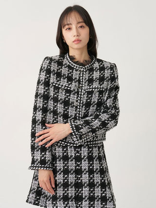 A woman wearing a black and white tweed houndstooth jacket with a high round collar and long sleeves, posed against a solid light gray background.