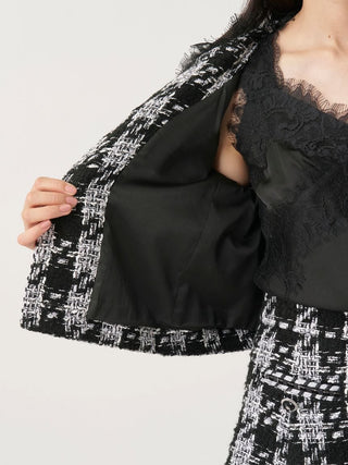 Close-up of a person wearing a black and white tweed jacket with wide flared sleeves, layered over a black lace camisole, against a plain light background.