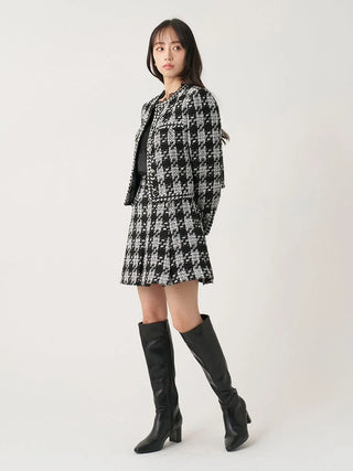 A woman modeling a black and white houndstooth tweed jacket and matching mini skirt set, paired with black knee-high leather boots, against a plain light gray studio background.
