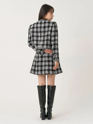 A woman shown from the back wearing a black and white houndstooth patterned mini dress with long sleeves and a tie belt, paired with black knee-high boots, against a plain light background.