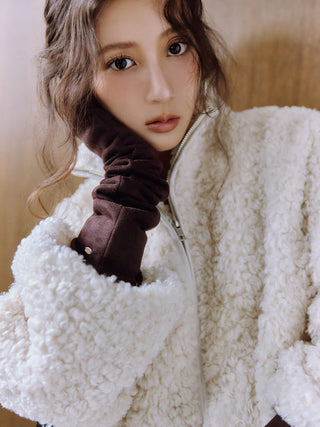 A close-up portrait of a woman wearing a fluffy white sherpa jacket and a dark brown ruched sleeveless glove, posed against a soft beige background.