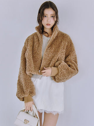 A woman models a cropped, light brown curly faux-fur zip-up jacket over a white top and white skirt, holding a small white handbag against a plain light gray studio background.