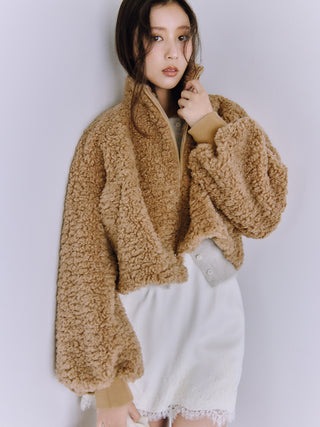A woman wearing a cropped, tan sherpa fleece zip-up jacket with ribbed cuffs and collar, paired with a white skirt, posing against a light gray studio background.