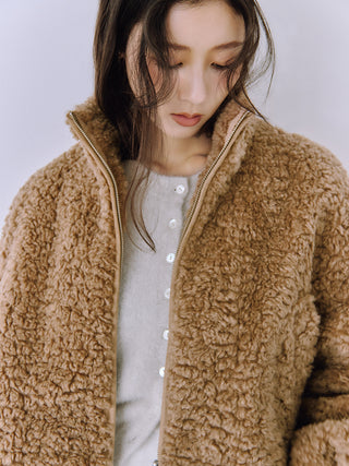 A woman wearing a warm camel brown sherpa fleece zip-up jacket over a light gray button-up top, photographed against a soft light gray background.