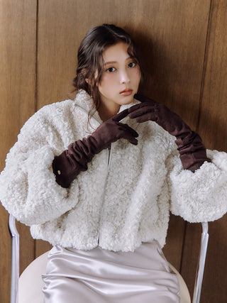 A woman wearing a fluffy cream sherpa cropped jacket, dark brown long gloves, and a shiny light gray skirt, posing against a wooden panel background.