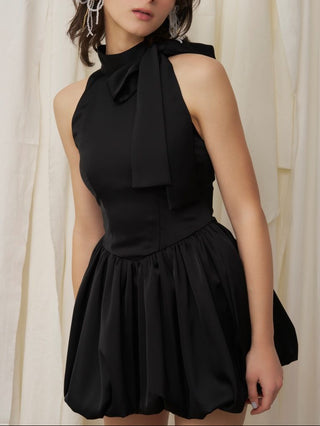 Woman wearing a black dress with a bow tie neckline against a white curtain background