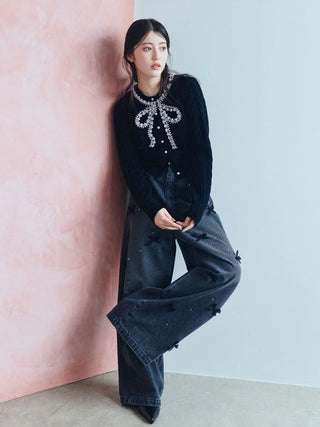 A woman leans against a pink wall wearing a black long-sleeve knit top with a sparkling rhinestone bow detail at the collar, paired with dark wide-leg jeans with star embellishments.