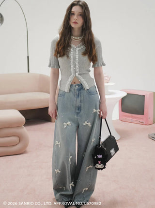 A model stands in a pink room wearing a gray short-sleeve knit top with white lace trim and light blue wide-leg denim jeans with bow embroidery. She is holding a black Sanrio bag with character details.