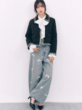A model stands against a plain white wall wearing a black quilted jacket with silver trim, a white ruffled blouse, and wide-leg light blue jeans with white embroidered details.