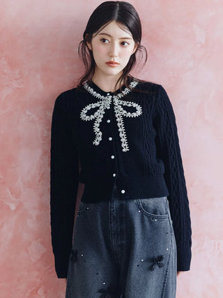 A woman wearing a black cable-knit cardigan with a white pearl bow detail at the collar, paired with gray denim jeans, standing against a textured pink wall.