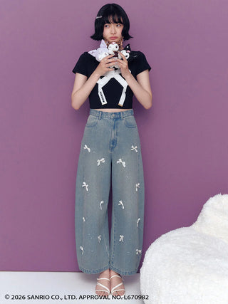 A model stands against a solid purple wall wearing a black short-sleeve crop top with a white bow graphic, light-wash wide-leg denim jeans with small white bow prints, and white strappy heels. She is holding a plush toy with a white bow.