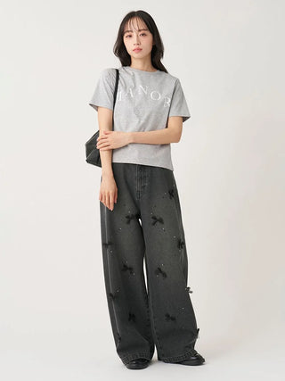 A model stands against a plain light background wearing a light gray short-sleeve t-shirt with white lettering, dark gray wide-leg jeans with black bow details, and black shoes.