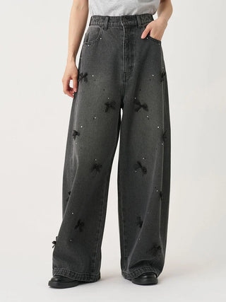 A person wearing wide-leg charcoal gray jeans with black bow and bead embellishments, paired with a light gray top and black shoes, photographed against a plain light background.