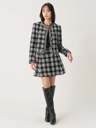 A model wearing a black and white houndstooth tweed jacket and matching mini skirt set with black leather knee-high boots, photographed against a plain light gray studio background.