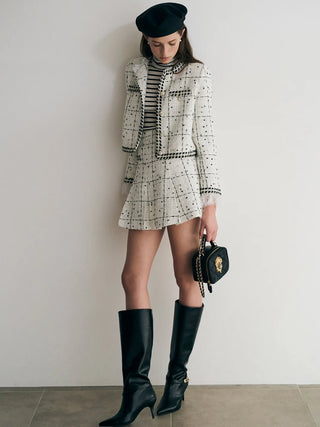 A model wearing a black and white tweed blazer and matching mini skirt set with a striped turtleneck, black leather knee-high boots, a black beret, and holding a small black handbag with a gold logo, standing against a plain off-white wall.