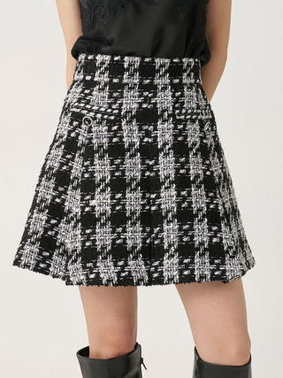 A black and white tweed mini skirt with a textured plaid pattern, worn by a model with a black top and black boots against a light gray background.