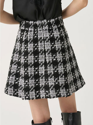 A black and white tweed plaid A-line skirt worn by a model, paired with a black satin top and black leather boots, shown from the waist down against a light gray studio background.