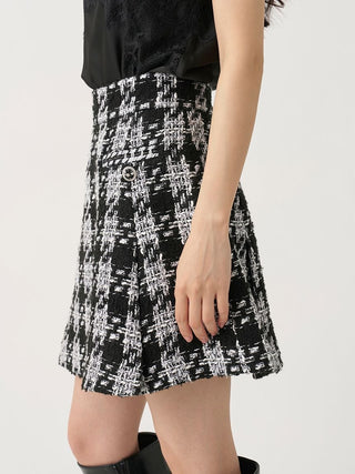 A close-up of a model wearing a black and white tweed mini skirt with a textured plaid pattern, paired with a black top and black boots against a plain light background.
