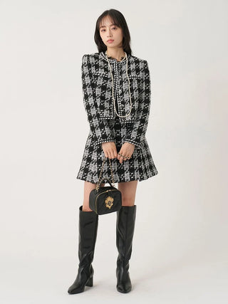 A model stands against a plain light background wearing a black-and-white tweed mini dress with a high collar, long sleeves, and a flared skirt. She is also wearing tall black knee-high boots and holding a small black handbag with a gold lion emblem.