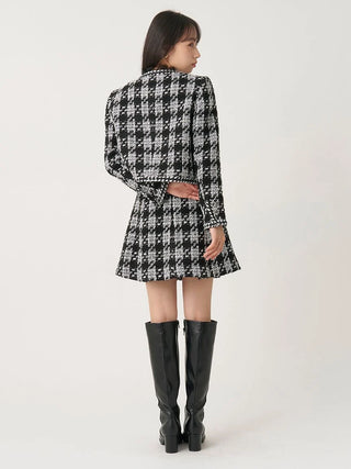 A woman shown from the back wearing a black and white houndstooth patterned mini dress with long sleeves and a tie waist, paired with black knee-high boots, against a plain light background.