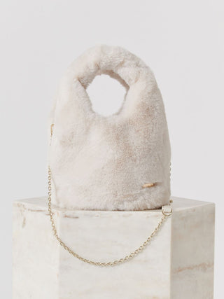 A fluffy faux-fur handbag in a light cream color with a gold chain strap, displayed on a white marble pedestal against a light gray background.