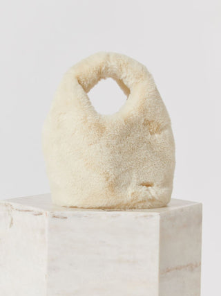 A cream-colored fluffy faux fur handbag with a top handle, displayed on a light marble pedestal against a light gray background.