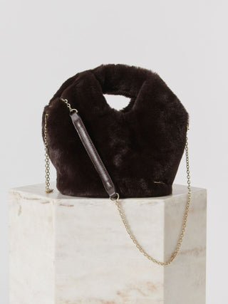 A dark brown faux fur shoulder bag with a gold chain strap, displayed on a white marble pedestal against a light gray background.