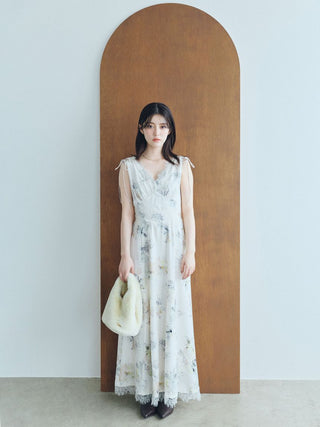 A woman stands in front of a large arched wooden panel wearing a sleeveless white maxi dress with a delicate blue and green floral print, holding a cream-colored fuzzy handbag.