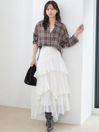 A woman stands indoors against a white wall corner wearing a brown plaid cropped button-up shirt, a long white tiered pleated skirt, and dark gray ankle boots, while holding a black handbag.